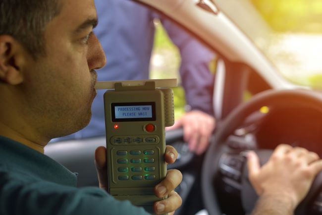 a man being pulled over and blowing into a breathalyzer in his car while a police officer watches 