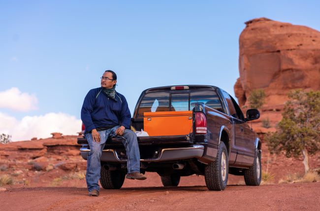 a man in a blue jacket and jeans sits in the bed of his pickup truck in the desert