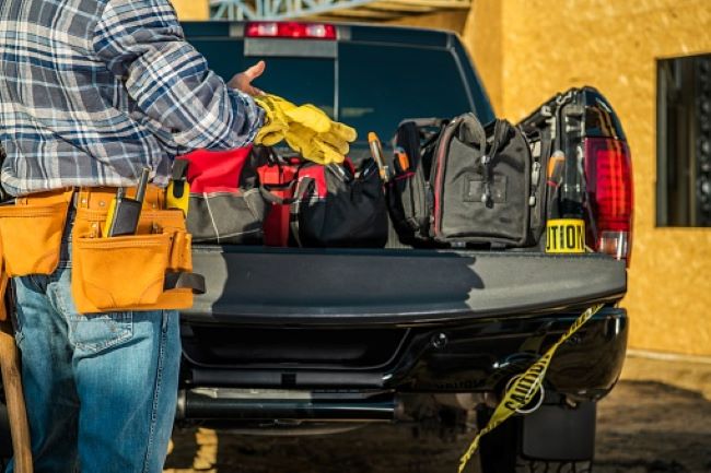 a man in a plaid shirt and work belt takes off his work gloves while looking at the tools in his work truck