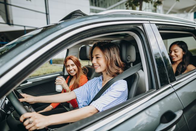 three girls drive in a black care while drinking coffee and laughing