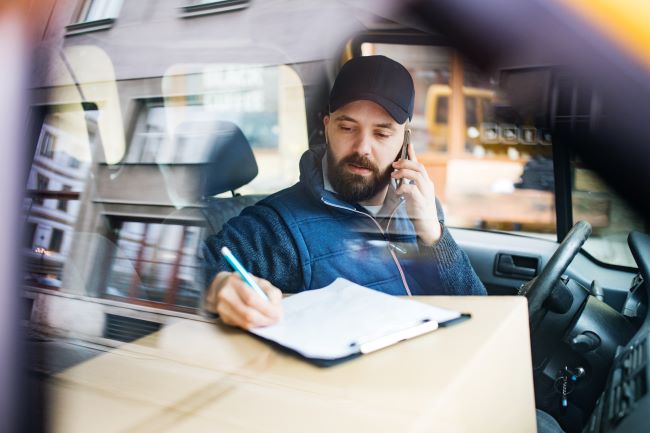 a man in a blue jacket and baseball cap talks on the phone while writing on a clipboard next to him