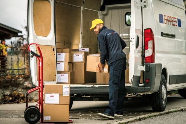 a man in a blue jumpsuit loads boxes into delivery service van from a red dolley